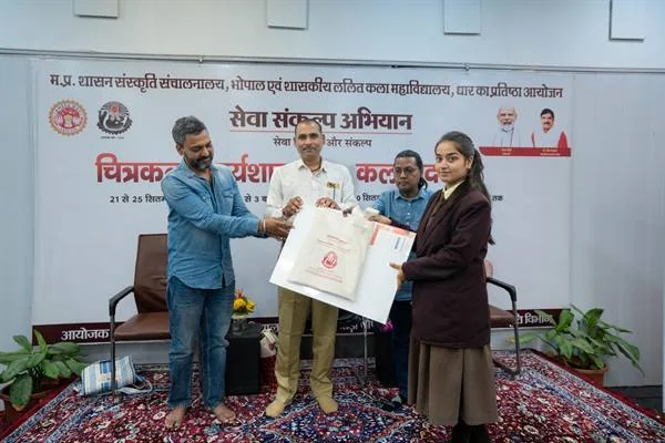 मंच पर तीन पुरुषों की मौजूदगी में एक युवती को कला सामग्री का पैकेट देते हुए अतिथि