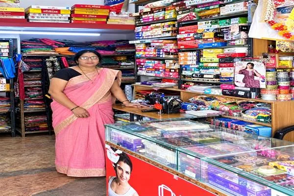 कपड़ों और सामान से भरी दुकान में गुलाबी साड़ी पहने महिला काउंटर के पास खड़ी हैं