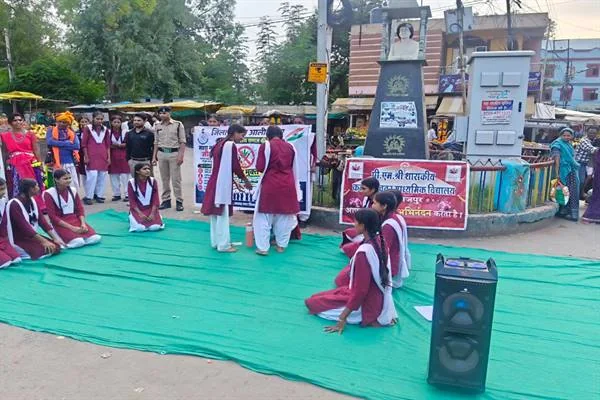 हरे कालीन पर मरून-सफेद वर्दी में नुक्कड़ नाटक प्रस्तुत करती छात्राएं, पास खड़े शिक्षक, पुलिसकर्मी और नागरिक
