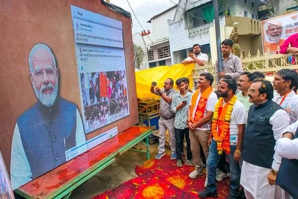 सड़क किनारे लगे बड़े होर्डिंग पर प्रधानमंत्री नरेंद्र मोदी की तस्वीर और एक सोशल मीडिया पोस्ट का स्क्रीनशॉट, पास में गेंदे की माला पहने कुछ लोग खड़े होकर देख रहे हैं और एक व्यक्ति मोबाइल से वीडियो बना रहा है।