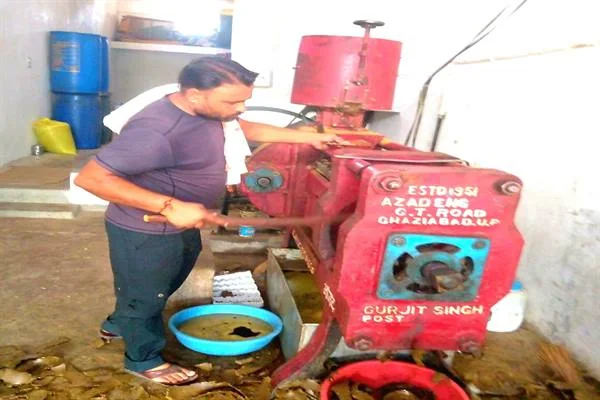 एक युवक कमरे में लगी लाल रंग की तेल पेराई मशीन के पास खड़े होकर उसे चला रहा है, मशीन के नीचे रखे बर्तन में तेल इकट्ठा हो रहा है।