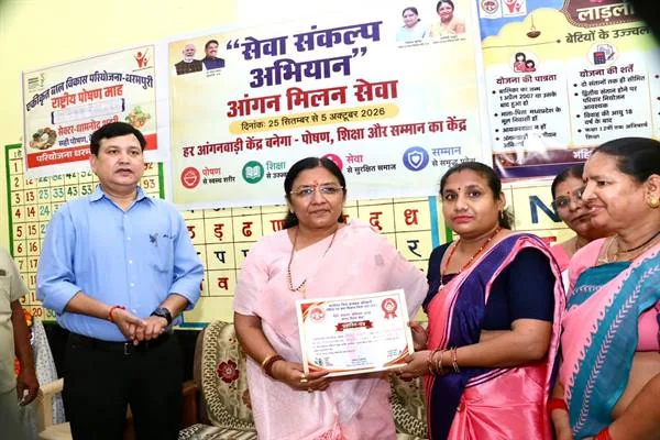 आंगनवाड़ी केन्द्र के भीतर एक महिला जनप्रतिनिधि दूसरी महिला को प्रमाण पत्र सौंपती हुईं, साथ में एक अधिकारी और दो अन्य महिलाएं खड़ी हैं; पीछे 'सेवा संकल्प अभियान – आंगन मिलन सेवा' का बैनर और दीवार पर हिंदी वर्णमाला व योजना संबंधी चार्ट लगे हैं।